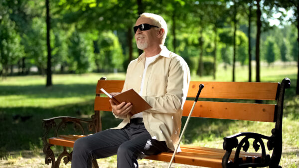 Happy blind person reading Braille on park bench, with cane next to him