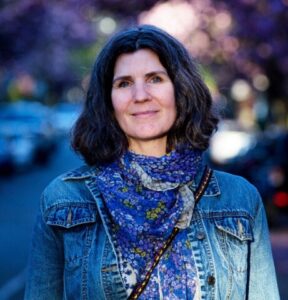 Headshot of Robbie McDonald, a cis-gendered white woman who is 55 years old, deaf, and neurodivergent. She has a welcoming expression with blue eyes while wearing a jean jacket and a scarf with blue flowers. She is standing on a street festooned with cherry blossoms.