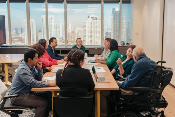 A group of diverse professionals representing various disability identities gather in a meeting around a conference table. Windows overlook a city skyline.