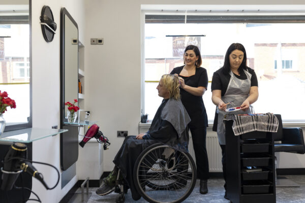 Side view of a man in a wheelchair visiting the hairdressers in the North East of England. The hairdressers are braiding his hair while also dying the ends of it with different colours.