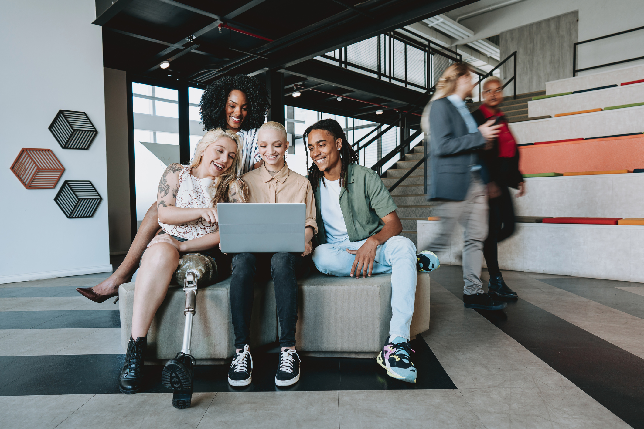 A group of four people sit together on a bench. The person in the middle is holding a laptop that the others are all looking at the screen of. Two more people walk past them in the background.