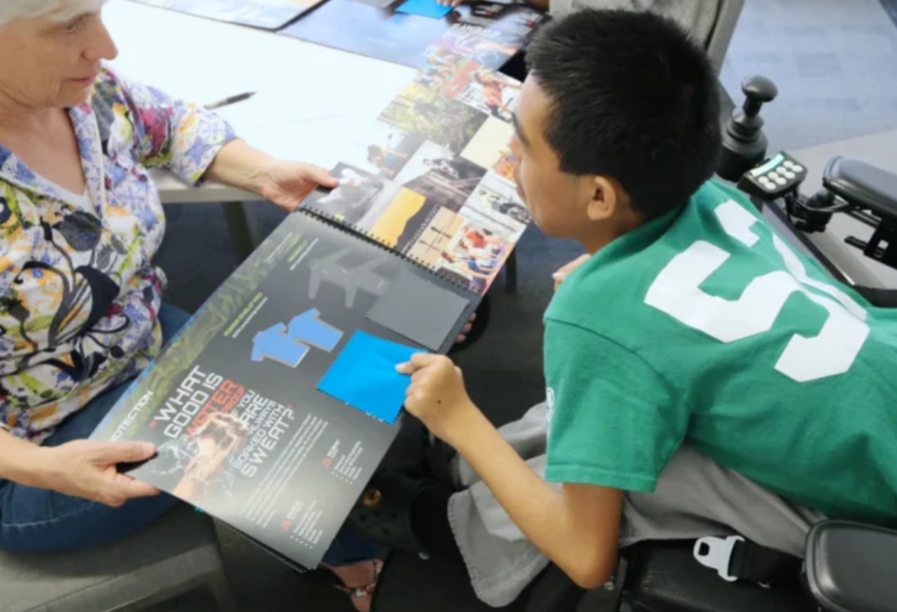 A motorized wheelchair user in a green shirt is pictured from the side. They are leaning forward to point at a flipbook being held open and presented to them by a person in a floral blouse.