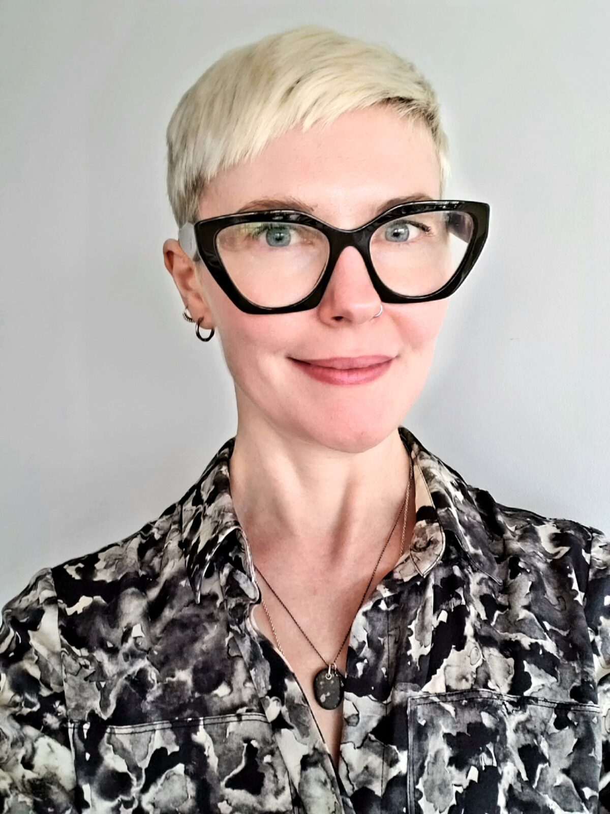Headshot for Mette, a smiling, femme-presenting woman in her 40s. She has short, very light hair, blue eyes, and is white-skinned. She is wearing large black glasses and a collared shirt with a black and grey abstract print.