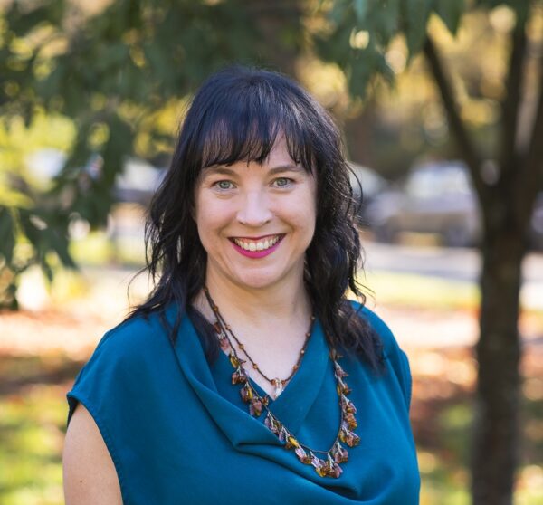 Headshot of Trish Kelly: In the foreground stands Trish Kelly, a light skinned woman with dark hair and thick black glasses. In the background is a park setting.