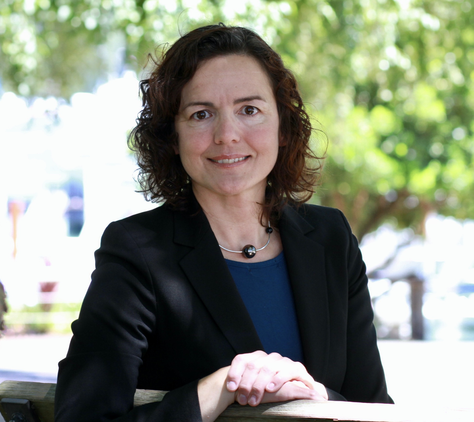 Headshot of Therese Boullard: She is a white woman with shoulder-length curly brown hair and brown eyes. She is wearing a black blazer with a blue shirt. She is outside in a park-like setting.