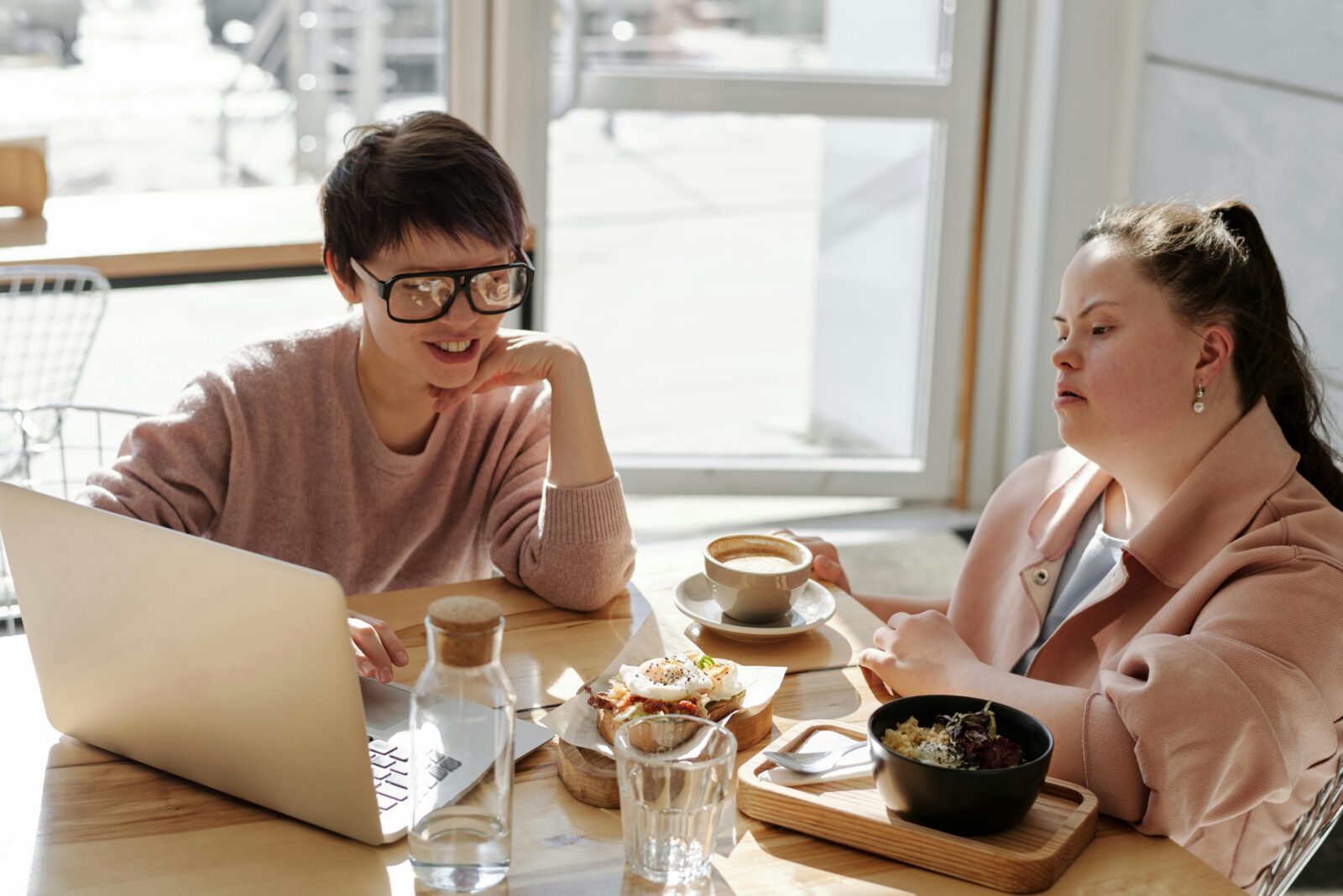 Two disabled people sitting at a table enjoying brunch while one of them is showing the other something on a laptop.