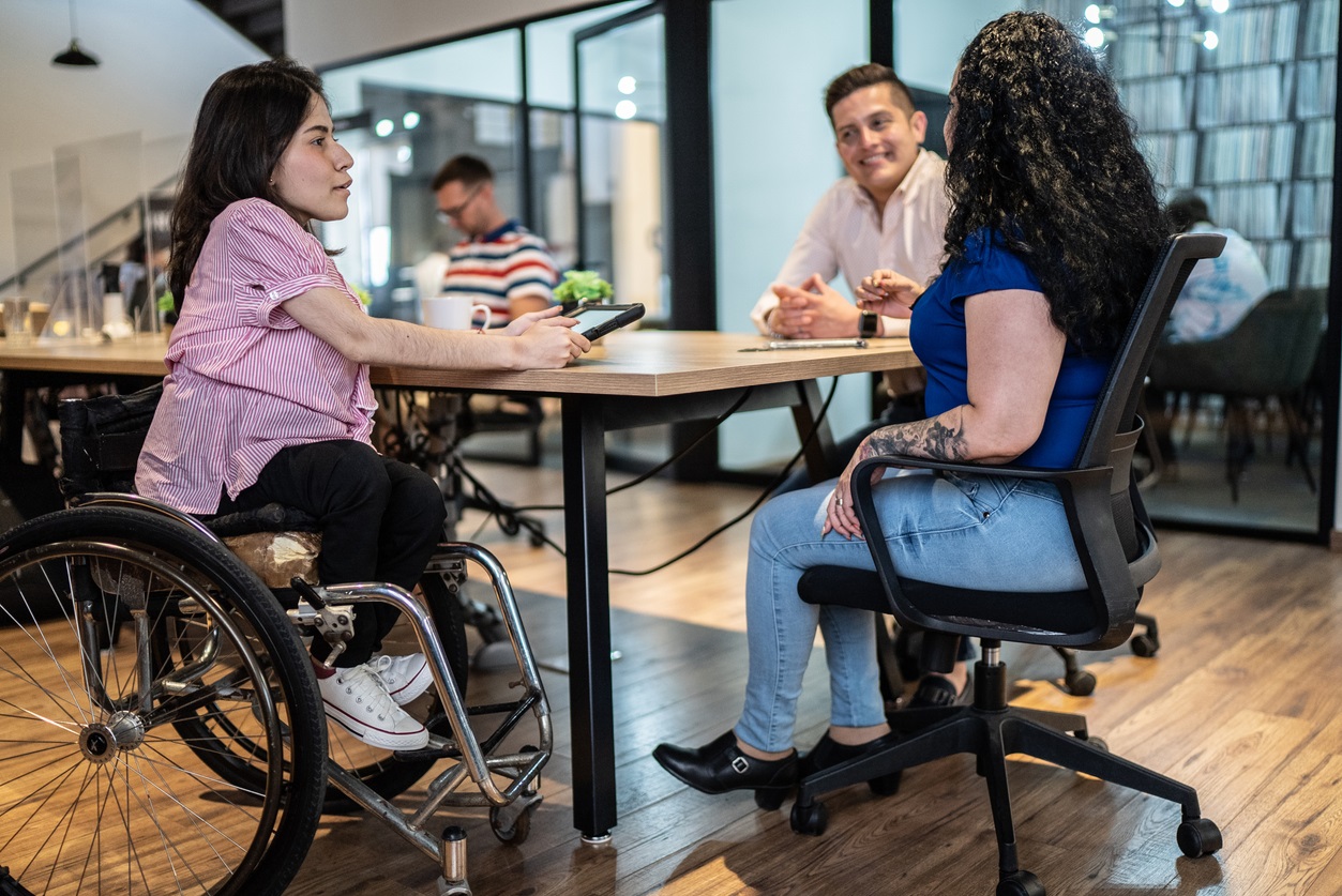 A group of coworkers meeting at a table. The woman in the foreground is a wheelchair user.